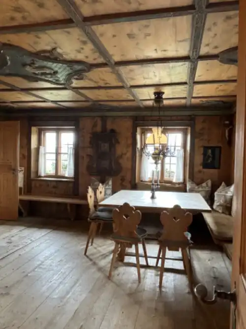 In the traditional dining room of the Hochalpschwendt Mountain Villa, there is a large wooden table, carved chairs, and a bench in front of the windows.