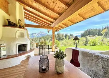 Auf der überdachten Chalet-Terrasse stehen Holzdecke, Kamin und Esstisch mit Blick auf Wiese, Wald und schneebedeckte Berge im Hintergrund.