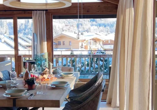In einem hellen Chalet von BelleStay steht ein gedeckter Essbereich vor großen Fenstern mit Blick auf verschneite Berge und Dächer.