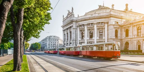 Bei Sonnenschein fährt eine rote Wiener Straßenbahn am Burgtheater vorbei entlang der Wiener Ringstraße durch das Zentrum von Wien.