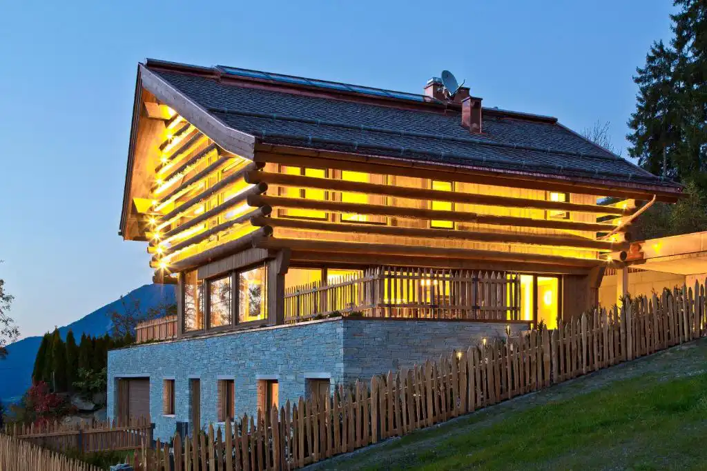 Das warm beleuchtete Holzferienhaus liegt am Hang und zeigt alpine Architektur mit steinernem Sockel, Zaun und Blick auf den Abendhimmel in den Bergen.