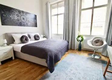 Das helle Schlafzimmer zeigt ein breites Bett mit dunkler Decke, zwei Kissen, Holzboden, große Fenster, Vorhänge, Teppich und eine kleine Sitzecke.